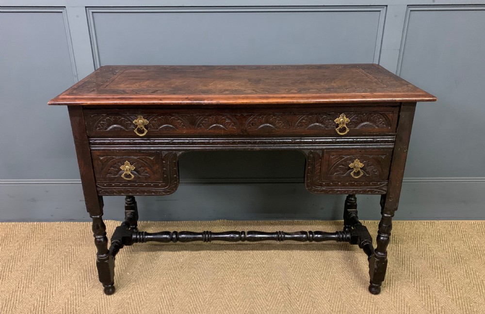 A dark carved oak side table featuring intricate lunette carvings, brass drop handles, three drawers, turned legs, and a stretcher base. View II