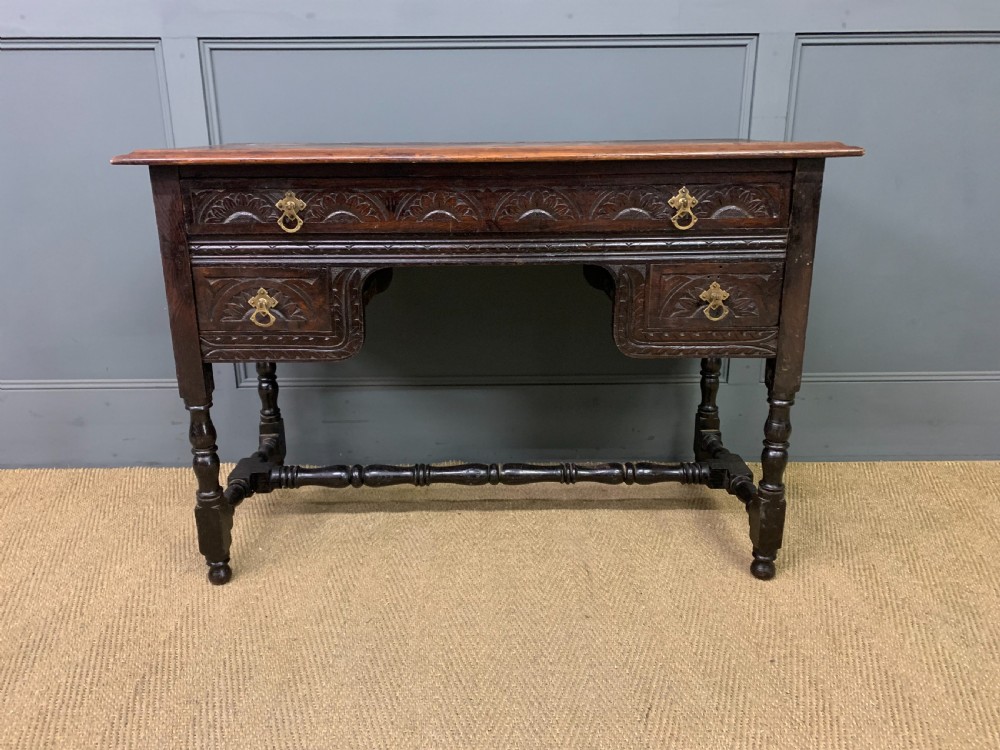 A dark carved oak side table featuring intricate lunette carvings, brass drop handles, three drawers, turned legs, and a stretcher base. View III