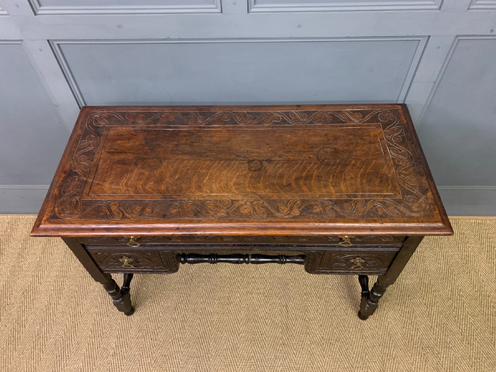 A dark carved oak side table featuring intricate lunette carvings, brass drop handles, three drawers, turned legs, and a stretcher base. View IV