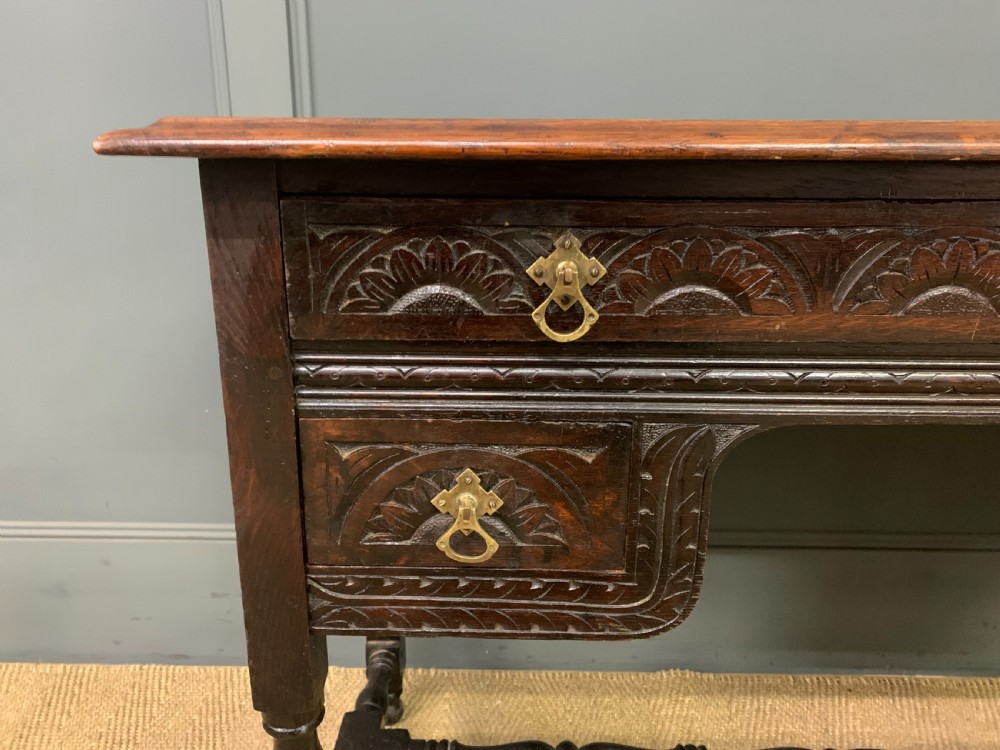 A dark carved oak side table featuring intricate lunette carvings, brass drop handles, three drawers, turned legs, and a stretcher base. View V