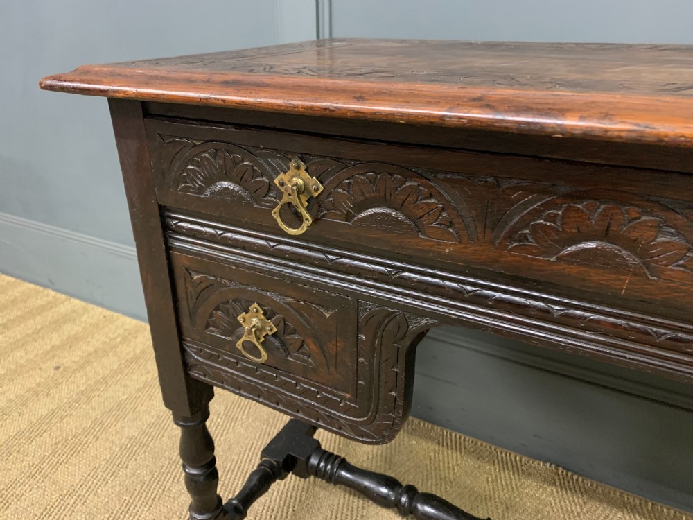 A dark carved oak side table featuring intricate lunette carvings, brass drop handles, three drawers, turned legs, and a stretcher base. View VI