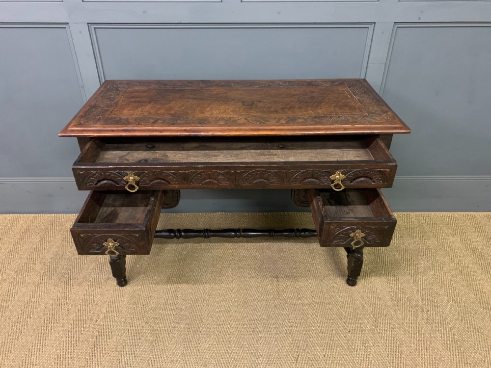 A dark carved oak side table featuring intricate lunette carvings, brass drop handles, three drawers, turned legs, and a stretcher base. View VIII