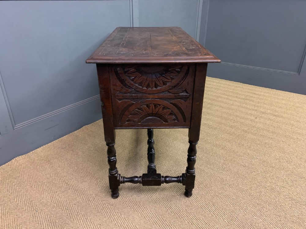 A dark carved oak side table featuring intricate lunette carvings, brass drop handles, three drawers, turned legs, and a stretcher base. View IX