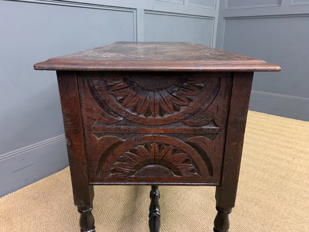 A dark carved oak side table featuring intricate lunette carvings, brass drop handles, three drawers, turned legs, and a stretcher base. View XI