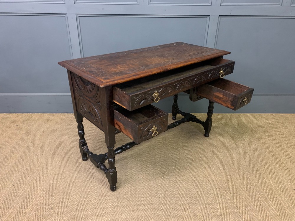 A dark carved oak side table featuring intricate lunette carvings, brass drop handles, three drawers, turned legs, and a stretcher base. View XII