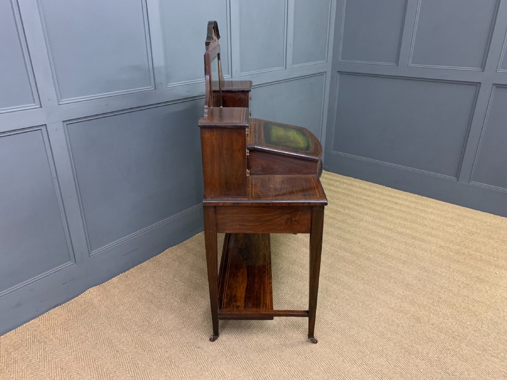 An antique rosewood bonheur du jour writing desk featuring intricate marquetry inlays, a central mirror, green leather writing top, and tapered legs on brass castors. View XVI