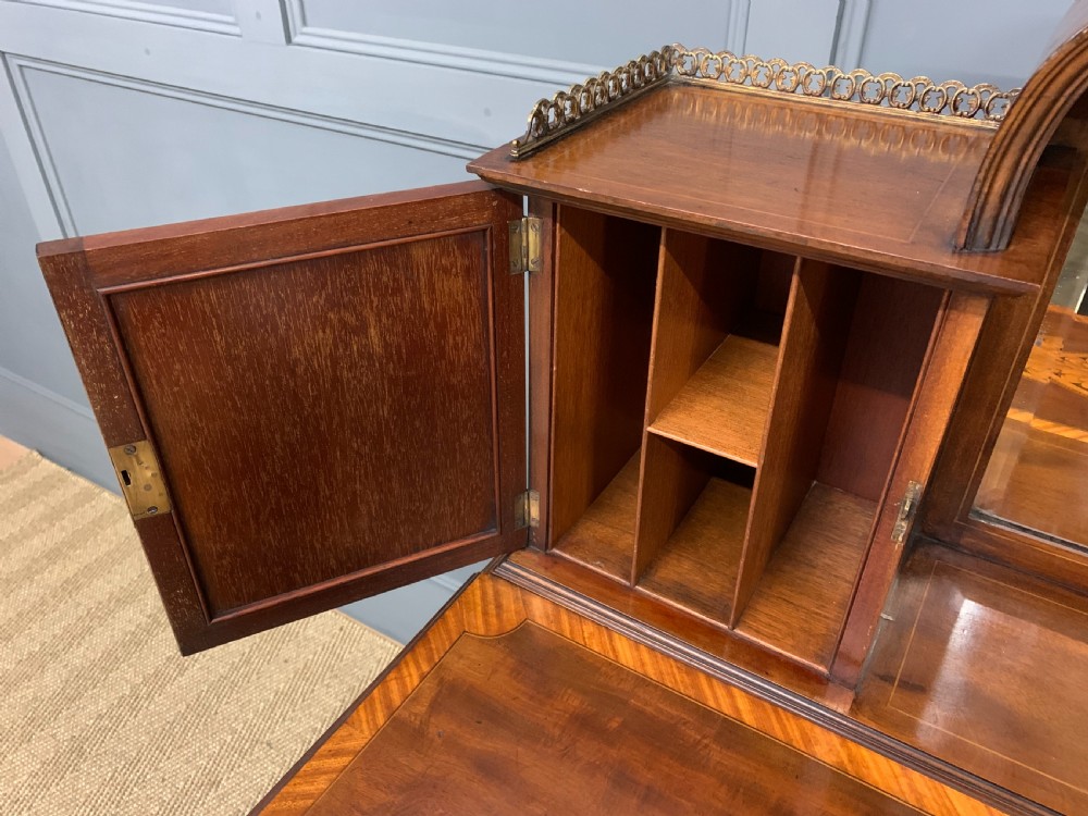 An Edwardian inlaid mahogany bonheur du jour writing desk featuring a central arched mirror, twin side cabinets with oval marquetry, brass galleries, and a lower shelf over tapered legs. View XVI