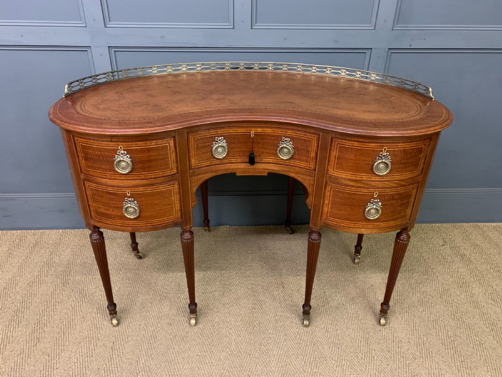 An Edwardian kidney-shaped mahogany writing desk featuring satinwood inlay, a leather writing surface, a brass gallery rail, and five drawers on fluted legs.