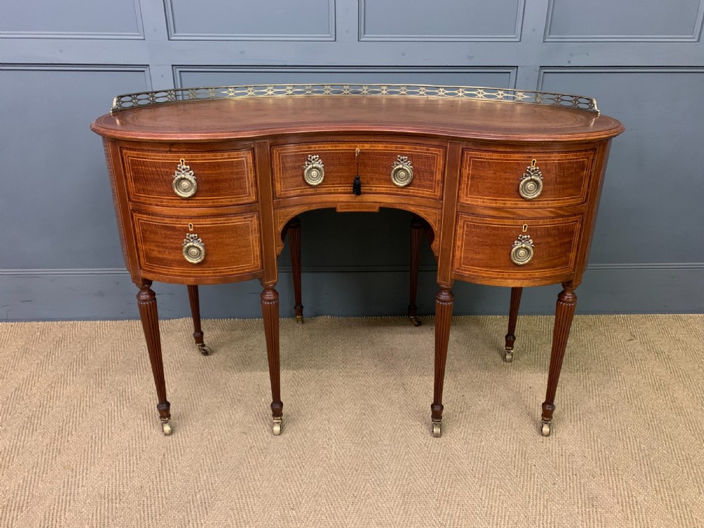 An Edwardian kidney-shaped mahogany writing desk featuring satinwood inlay, a leather writing surface, a brass gallery rail, and five drawers on fluted legs. View II