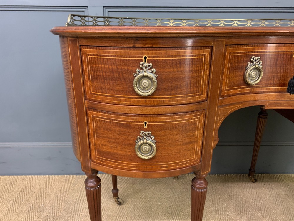 An Edwardian kidney-shaped mahogany writing desk featuring satinwood inlay, a leather writing surface, a brass gallery rail, and five drawers on fluted legs. View V