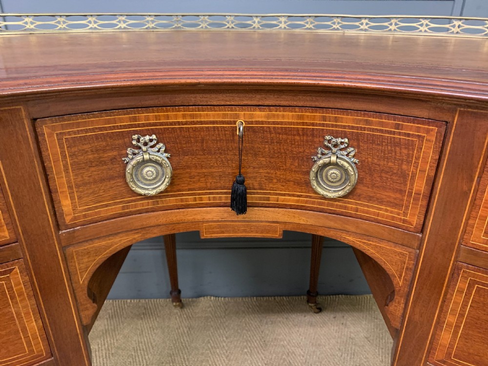 An Edwardian kidney-shaped mahogany writing desk featuring satinwood inlay, a leather writing surface, a brass gallery rail, and five drawers on fluted legs. View VI