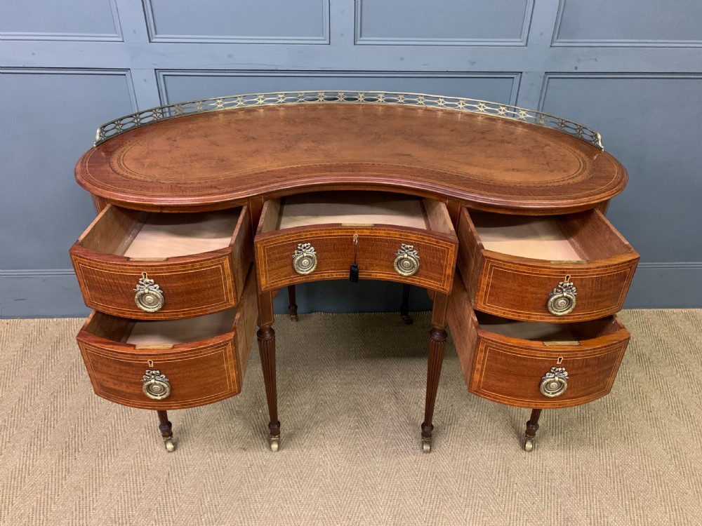 An Edwardian kidney-shaped mahogany writing desk featuring satinwood inlay, a leather writing surface, a brass gallery rail, and five drawers on fluted legs. View IX
