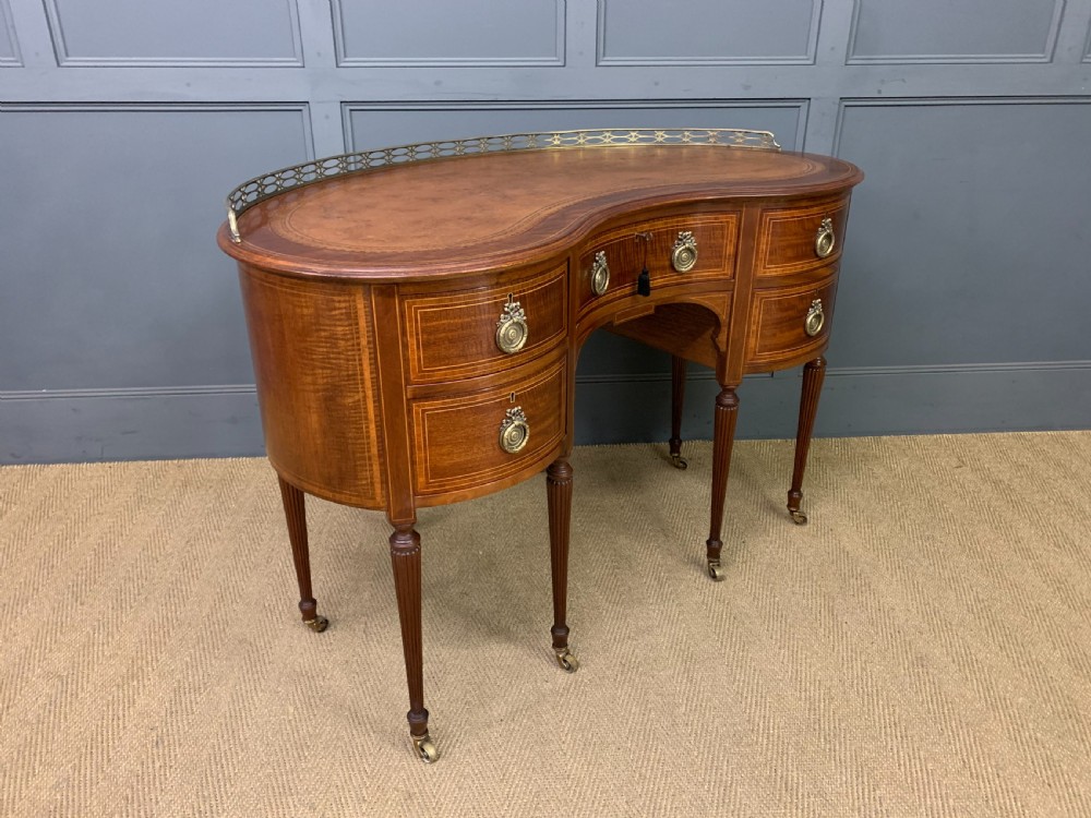 An Edwardian kidney-shaped mahogany writing desk featuring satinwood inlay, a leather writing surface, a brass gallery rail, and five drawers on fluted legs. View X