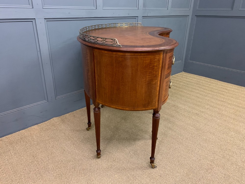 An Edwardian kidney-shaped mahogany writing desk featuring satinwood inlay, a leather writing surface, a brass gallery rail, and five drawers on fluted legs. View XI