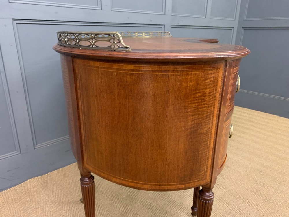 An Edwardian kidney-shaped mahogany writing desk featuring satinwood inlay, a leather writing surface, a brass gallery rail, and five drawers on fluted legs. View XII