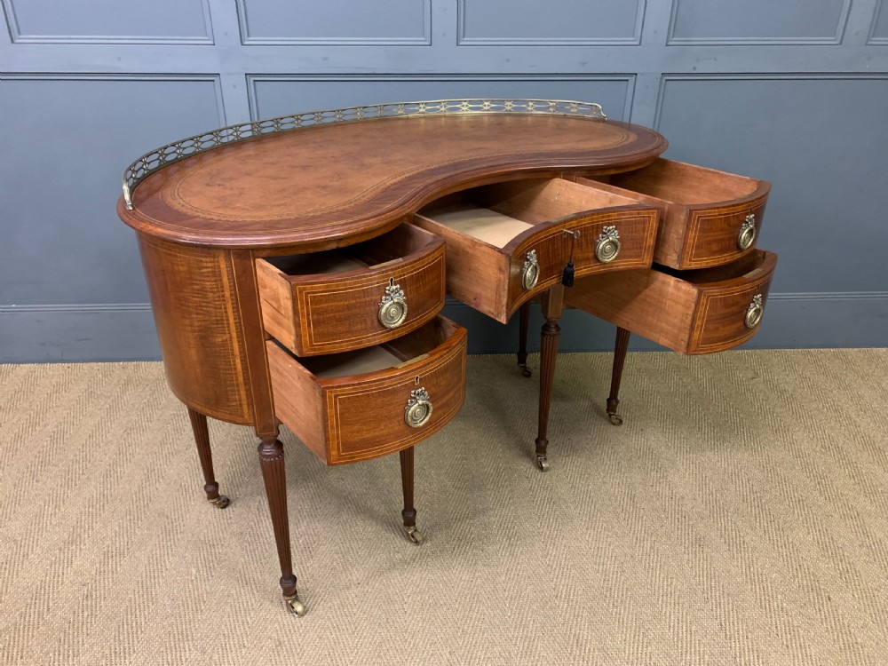 An Edwardian kidney-shaped mahogany writing desk featuring satinwood inlay, a leather writing surface, a brass gallery rail, and five drawers on fluted legs. View XIII