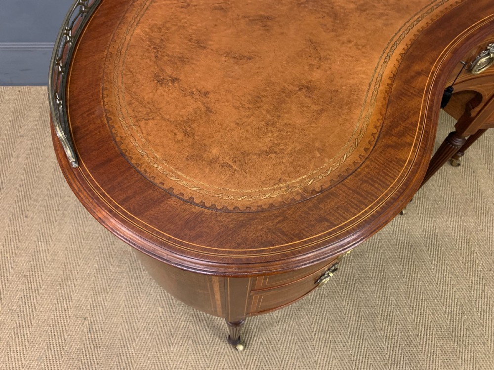 An Edwardian kidney-shaped mahogany writing desk featuring satinwood inlay, a leather writing surface, a brass gallery rail, and five drawers on fluted legs. View XIV