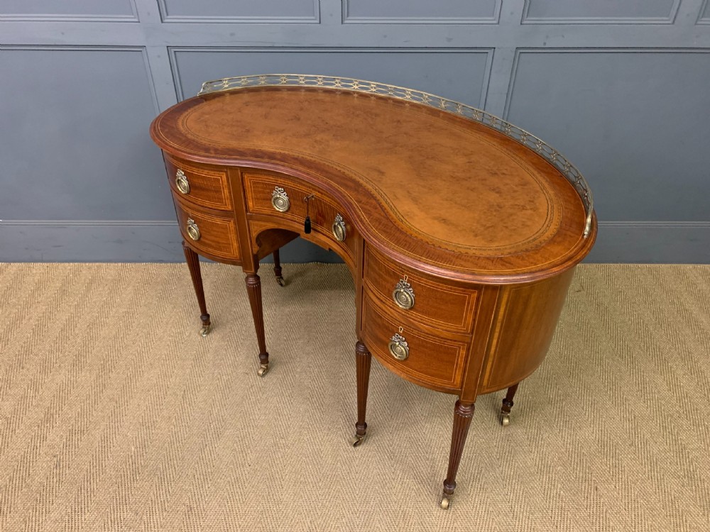 An Edwardian kidney-shaped mahogany writing desk featuring satinwood inlay, a leather writing surface, a brass gallery rail, and five drawers on fluted legs. View XV