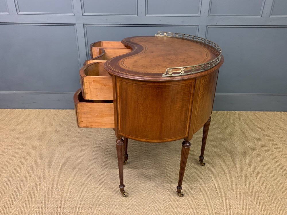 An Edwardian kidney-shaped mahogany writing desk featuring satinwood inlay, a leather writing surface, a brass gallery rail, and five drawers on fluted legs. View XVI