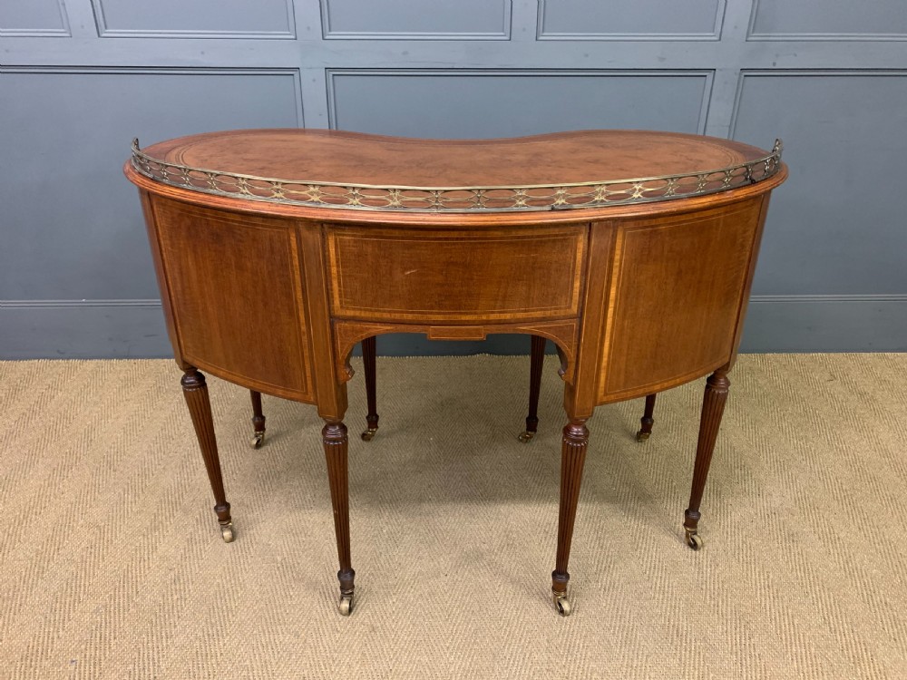 An Edwardian kidney-shaped mahogany writing desk featuring satinwood inlay, a leather writing surface, a brass gallery rail, and five drawers on fluted legs. View XVII