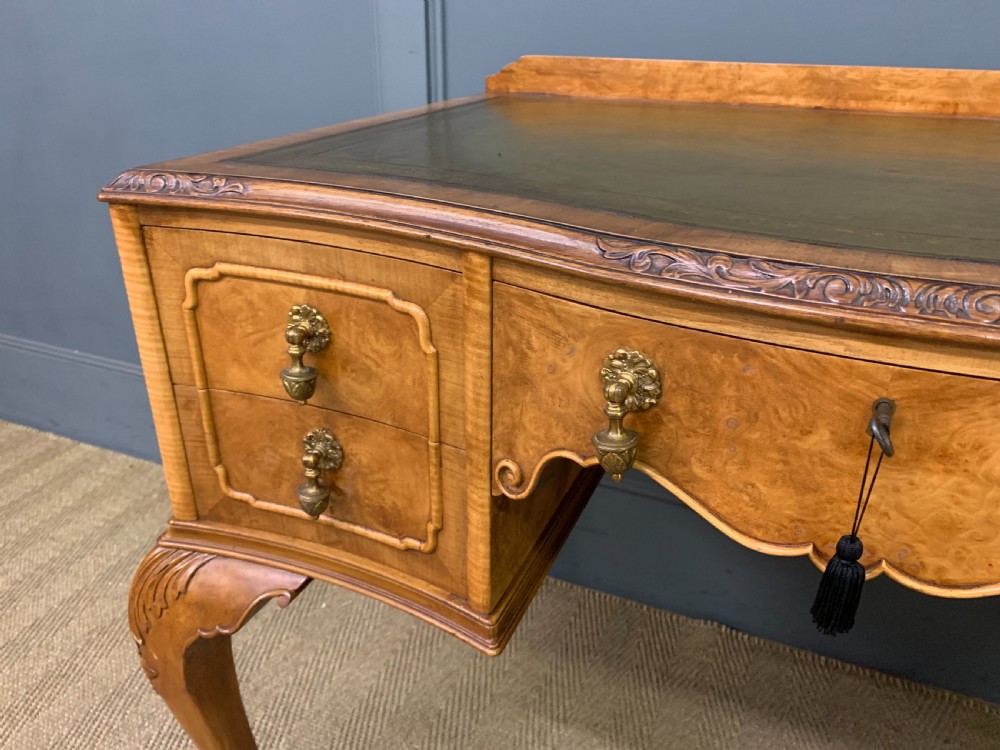 A Queen Anne style burr walnut writing desk with an inset green leather surface, five drawers, brass drop pulls, and carved cabriole legs. View VII