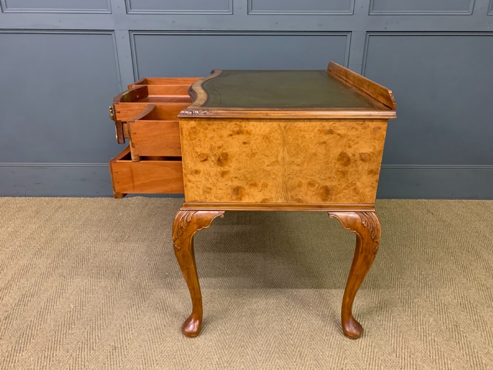 A Queen Anne style burr walnut writing desk with an inset green leather surface, five drawers, brass drop pulls, and carved cabriole legs. View XVI