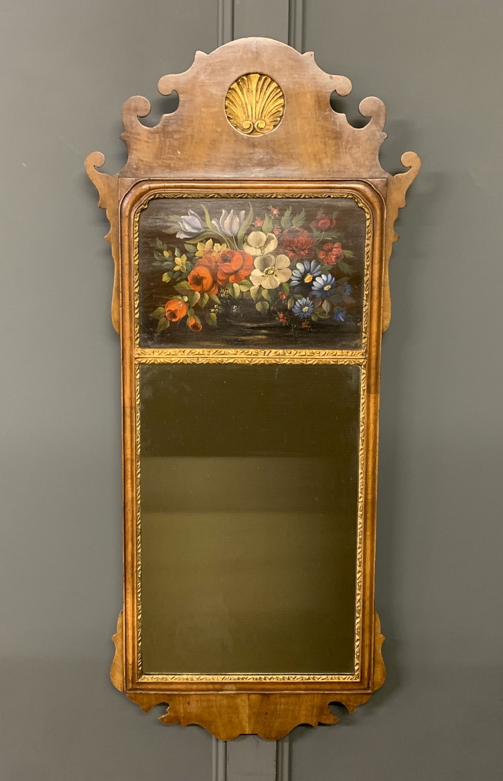 A vertical Queen Anne Revival walnut wall mirror featuring a scroll-cut frame, a gilded carved shell at the top crest, and a hand-painted floral still-life panel above the reflective mirror glass.