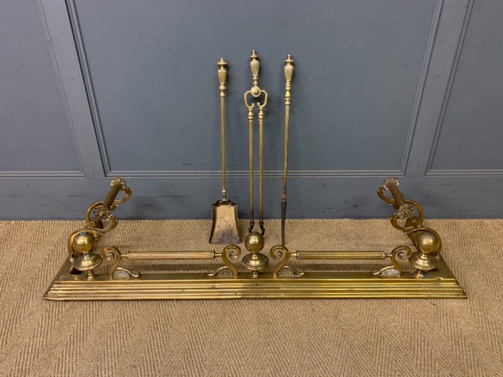 A Victorian brass fireplace fender featuring decorative finials and scrollwork rests, shown with three matching brass fire tools including a shovel, tongs, and poker.