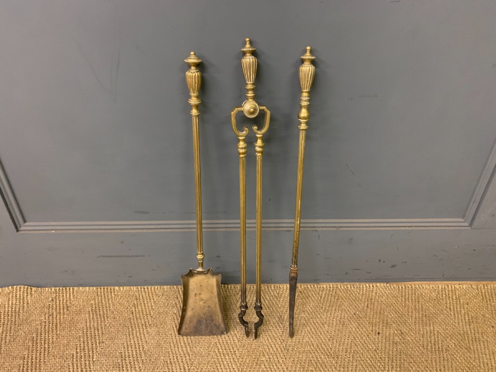 A Victorian brass fireplace fender featuring decorative finials and scrollwork rests, shown with three matching brass fire tools including a shovel, tongs, and poker. View II