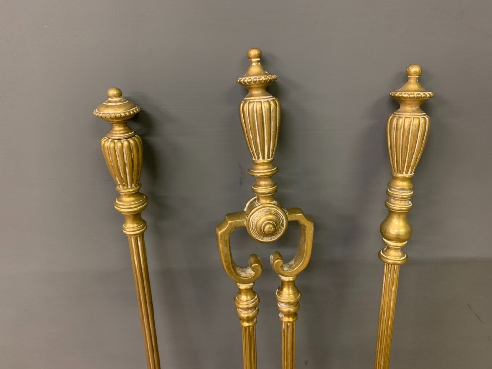 A Victorian brass fireplace fender featuring decorative finials and scrollwork rests, shown with three matching brass fire tools including a shovel, tongs, and poker. View III