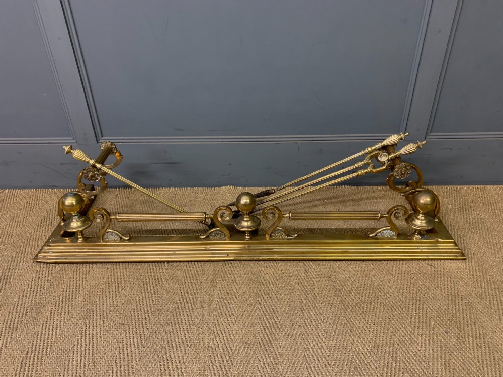A Victorian brass fireplace fender featuring decorative finials and scrollwork rests, shown with three matching brass fire tools including a shovel, tongs, and poker. View IV