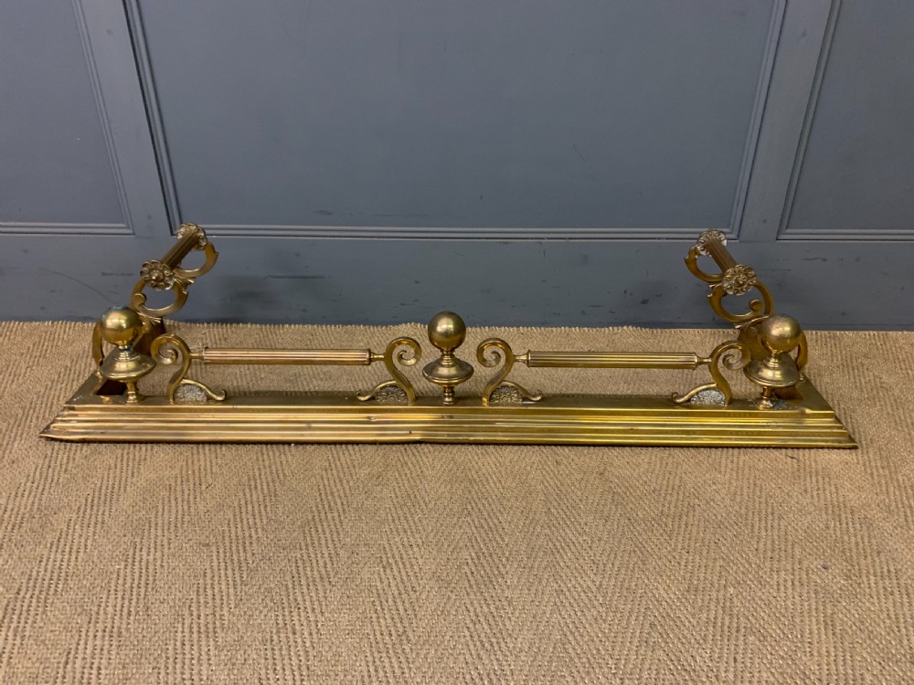 A Victorian brass fireplace fender featuring decorative finials and scrollwork rests, shown with three matching brass fire tools including a shovel, tongs, and poker. View V