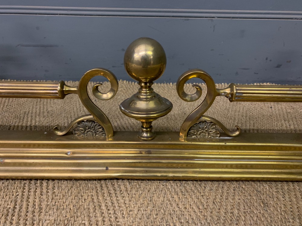 A Victorian brass fireplace fender featuring decorative finials and scrollwork rests, shown with three matching brass fire tools including a shovel, tongs, and poker. View VI