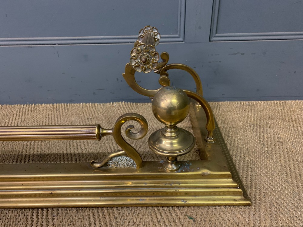 A Victorian brass fireplace fender featuring decorative finials and scrollwork rests, shown with three matching brass fire tools including a shovel, tongs, and poker. View VII