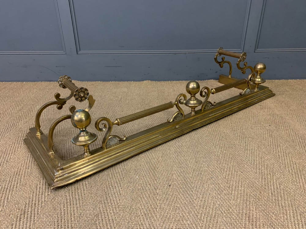 A Victorian brass fireplace fender featuring decorative finials and scrollwork rests, shown with three matching brass fire tools including a shovel, tongs, and poker. View IX