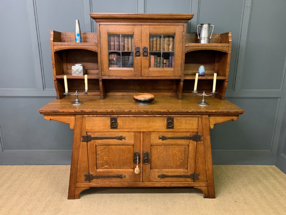 An Arts and Crafts period oak dresser featuring leaded glass upper cabinet doors, open side shelves, metal strap hinges, and a lower two-door cupboard.