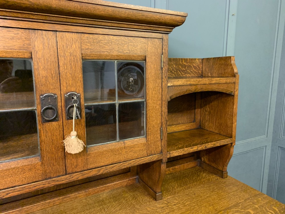 An Arts and Crafts period oak dresser featuring leaded glass upper cabinet doors, open side shelves, metal strap hinges, and a lower two-door cupboard. View VI