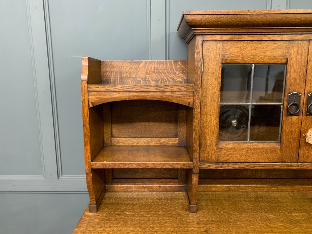 An Arts and Crafts period oak dresser featuring leaded glass upper cabinet doors, open side shelves, metal strap hinges, and a lower two-door cupboard. View VII