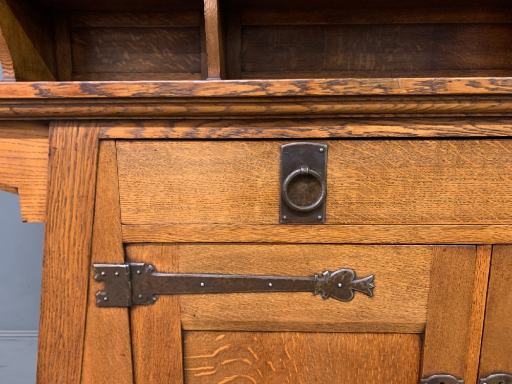An Arts and Crafts period oak dresser featuring leaded glass upper cabinet doors, open side shelves, metal strap hinges, and a lower two-door cupboard. View X