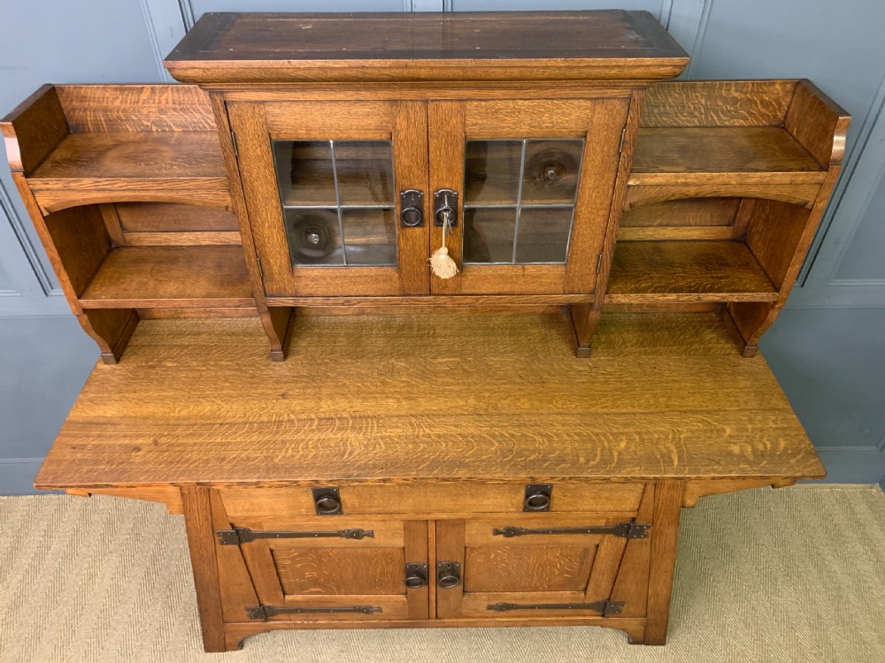 An Arts and Crafts period oak dresser featuring leaded glass upper cabinet doors, open side shelves, metal strap hinges, and a lower two-door cupboard. View XIV