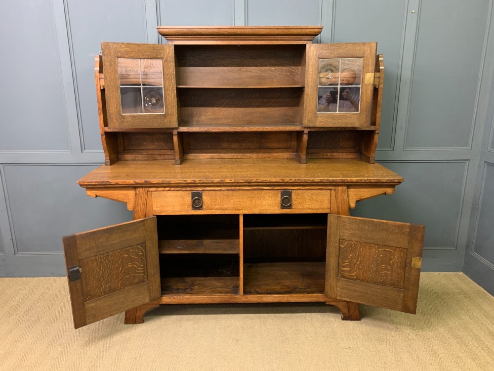 An Arts and Crafts period oak dresser featuring leaded glass upper cabinet doors, open side shelves, metal strap hinges, and a lower two-door cupboard. View XVI