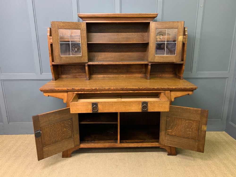 An Arts and Crafts period oak dresser featuring leaded glass upper cabinet doors, open side shelves, metal strap hinges, and a lower two-door cupboard. View XVII