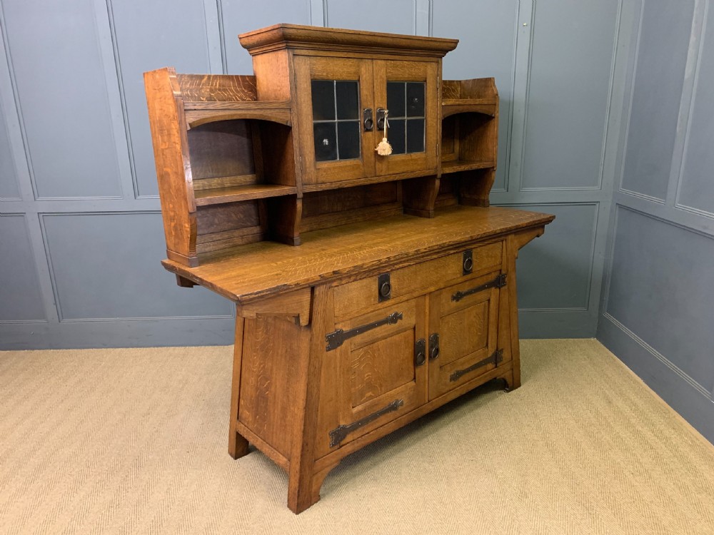 An Arts and Crafts period oak dresser featuring leaded glass upper cabinet doors, open side shelves, metal strap hinges, and a lower two-door cupboard. View XIX
