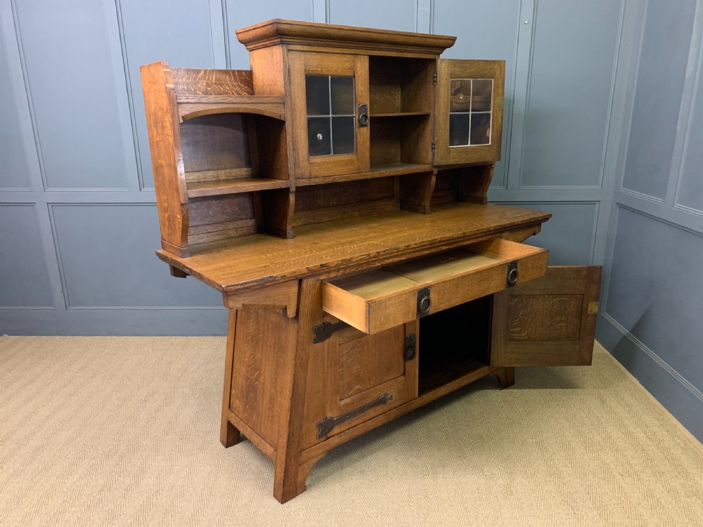 An Arts and Crafts period oak dresser featuring leaded glass upper cabinet doors, open side shelves, metal strap hinges, and a lower two-door cupboard. View XXII