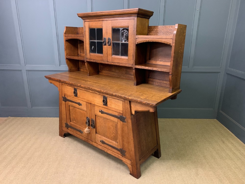 An Arts and Crafts period oak dresser featuring leaded glass upper cabinet doors, open side shelves, metal strap hinges, and a lower two-door cupboard. View XXIII