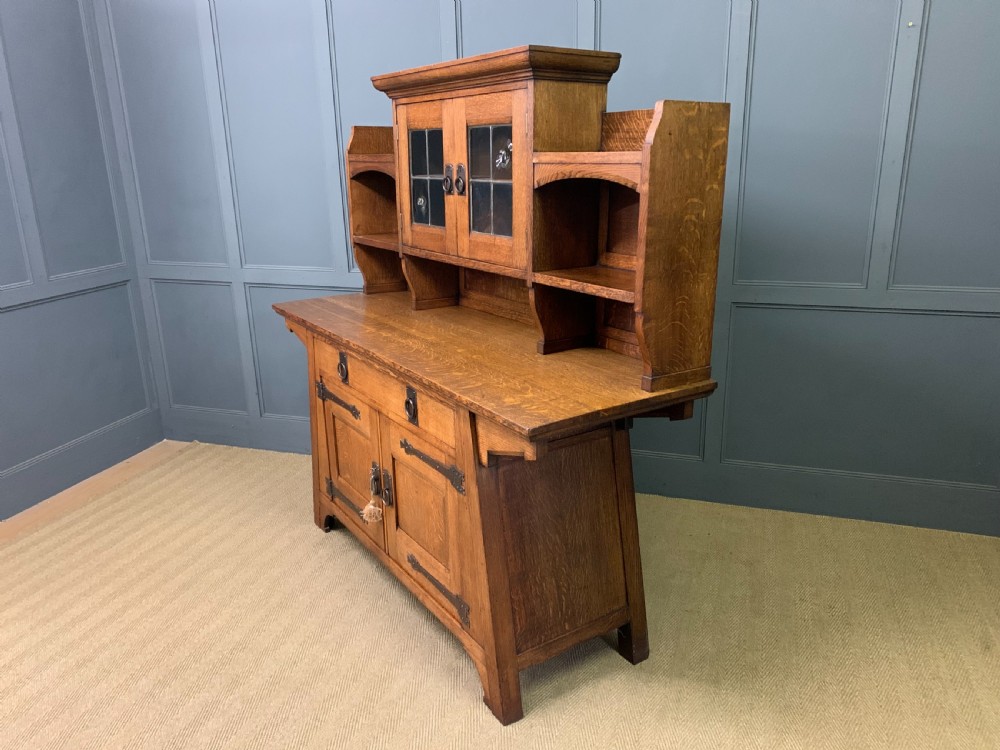 An Arts and Crafts period oak dresser featuring leaded glass upper cabinet doors, open side shelves, metal strap hinges, and a lower two-door cupboard. View XXIV