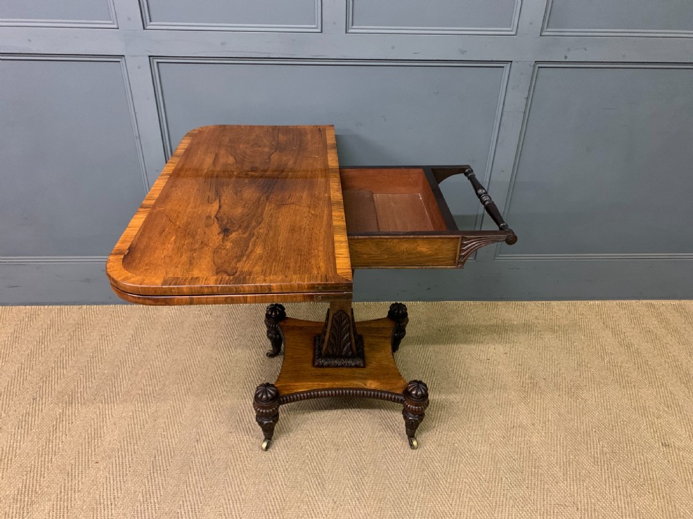 A Regency period rosewood fold-over card table with a swivel top, a central pedestal support, and a concave platform base resting on carved feet with brass castors. View X