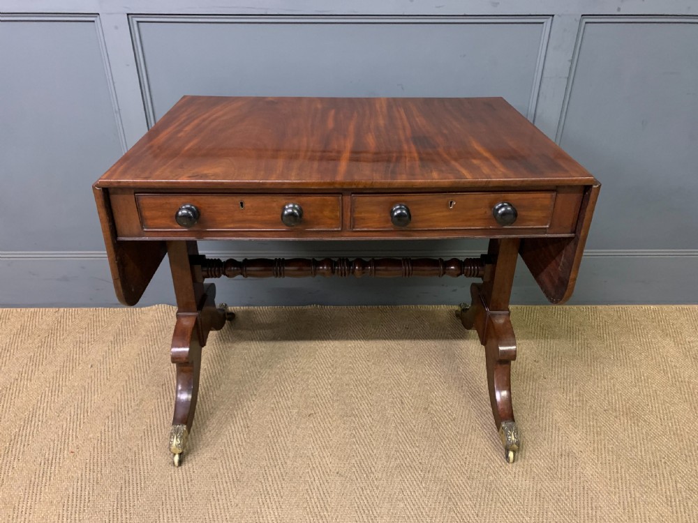 A George IV mahogany sofa table featuring two drawers with dark wooden knobs, drop-leaf side flaps, a turned central stretcher, and splayed legs on brass castors.