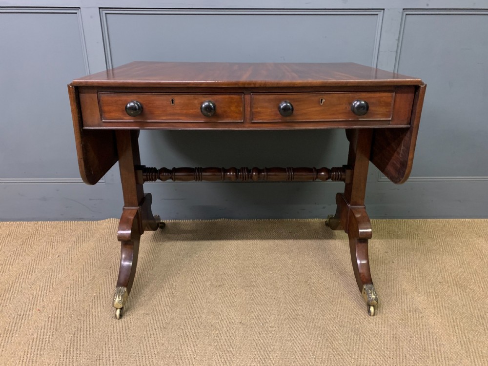 A George IV mahogany sofa table featuring two drawers with dark wooden knobs, drop-leaf side flaps, a turned central stretcher, and splayed legs on brass castors. View II
