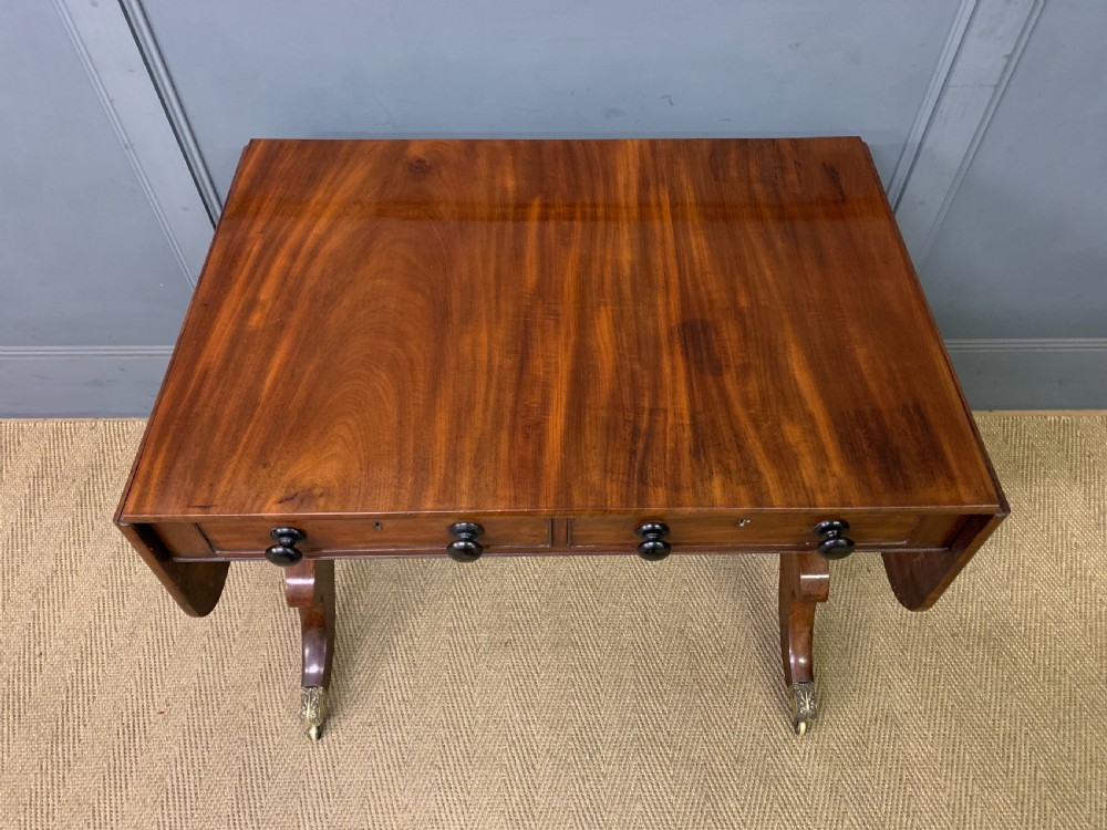 A George IV mahogany sofa table featuring two drawers with dark wooden knobs, drop-leaf side flaps, a turned central stretcher, and splayed legs on brass castors. View III